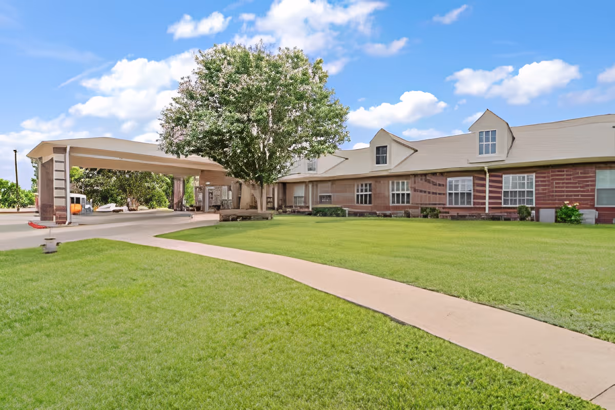 Exterior view of Morada Abilene facility showing a large green lawn, a curved concrete walkway, a tree near the entrance, and a building with multiple windows under a blue sky with scattered clouds.