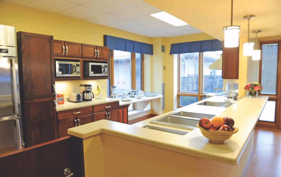 Bright, spacious kitchen with a long island and sink, a bowl of fruit on the counter, wooden cabinets, microwaves, and large windows.