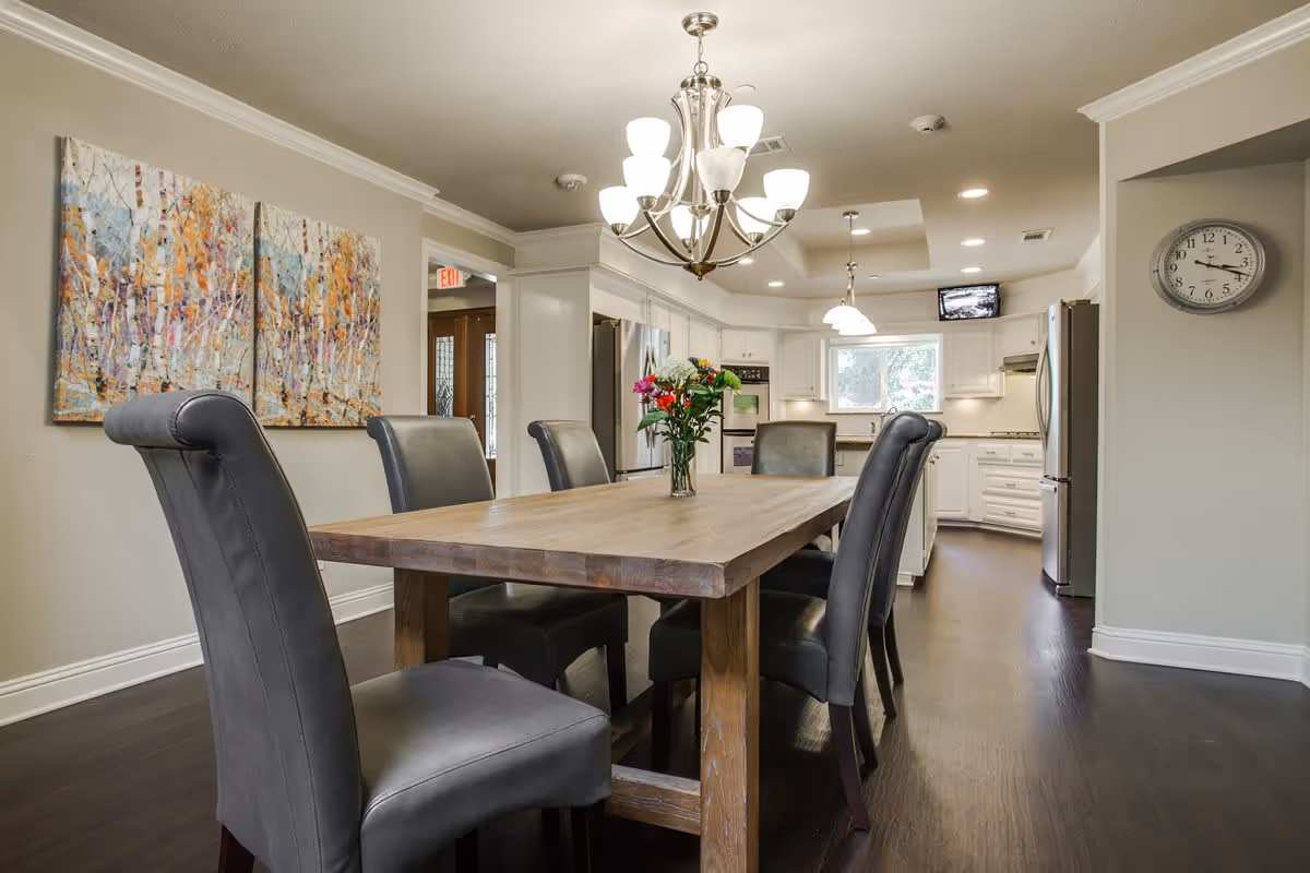 A modern dining area with a wooden table surrounded by six gray upholstered chairs. A vase with colorful flowers is placed in the center of the table. The room features a chandelier with multiple lights hanging above the table. In the background, there is a white kitchen with stainless steel appliances, including a refrigerator and oven. A wall clock is mounted on the right wall, and a colorful abstract painting hangs on the left wall.