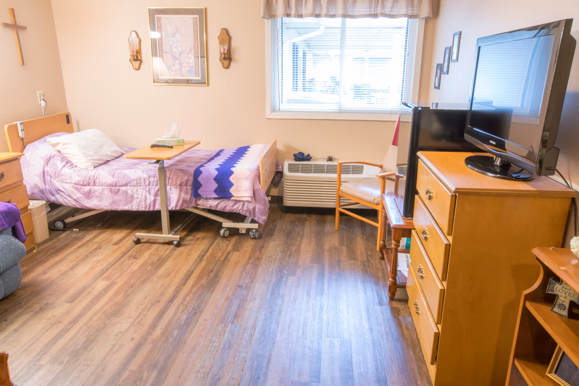 A senior living facility bedroom with a single bed covered in a purple blanket and a blue and purple crocheted throw. There is a small overbed table with a box of tissues on it. The room has wood flooring, a wooden dresser with a flat-screen TV on top, a wooden chair, and a window with blinds and a valance. The walls are decorated with a cross, a framed picture, and two wall sconces with candles.