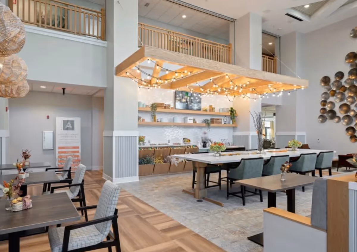 A bright and modern dining area with multiple tables and chairs. The space features a large wooden light fixture with string lights hanging above a long table with green upholstered chairs. There are decorative shelves on the wall with plants and other items, and a unique wall art installation made of circular metallic discs. The flooring is a mix of wood and tile, and the room has a high ceiling with a balcony railing visible above.