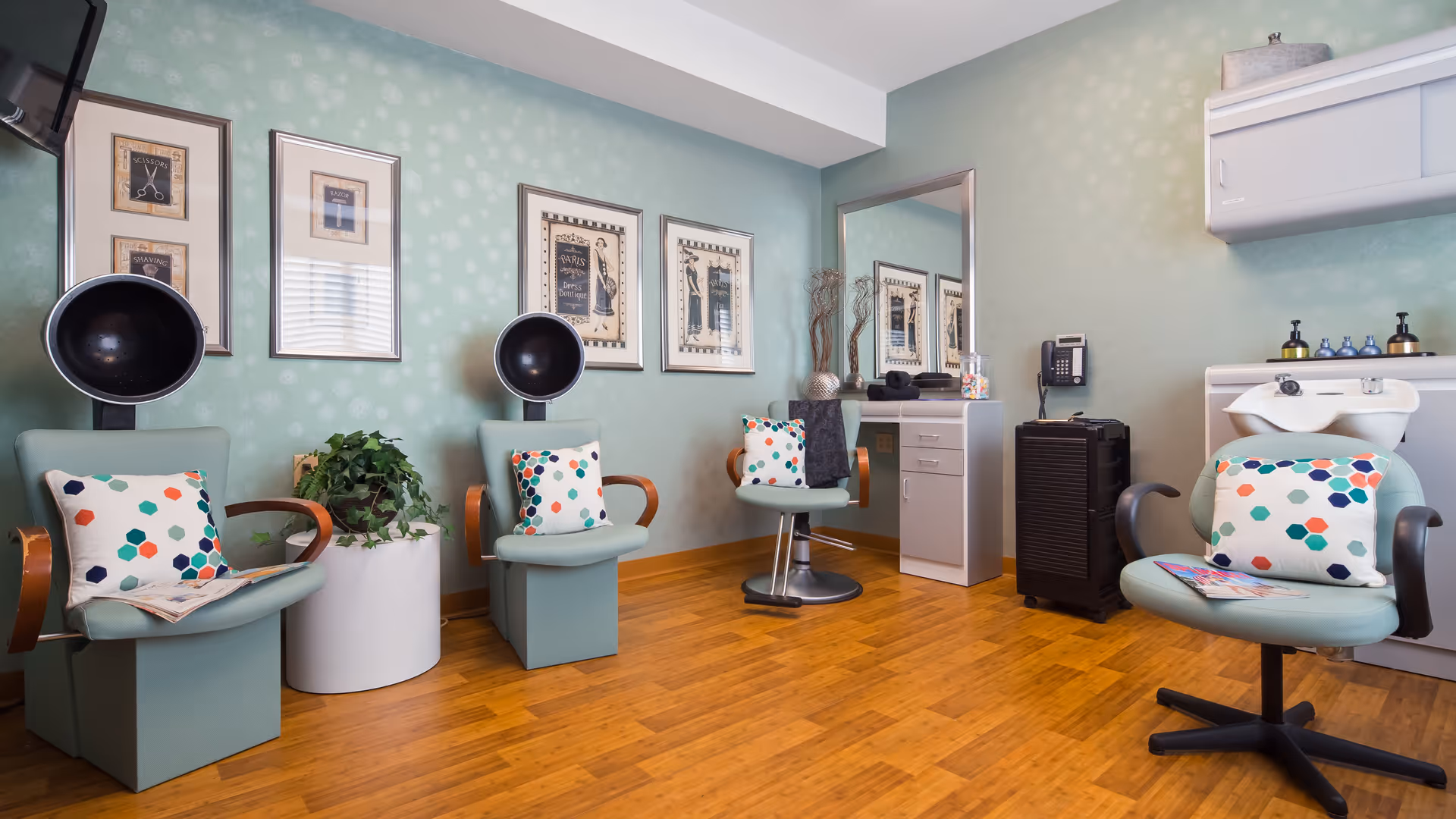 A bright and clean hair salon area in an assisted living facility featuring four vintage-style hair drying chairs with colorful polka dot pillows. The room has light green patterned walls adorned with framed artwork, a large mirror above a white vanity with drawers, a black rolling cart, and a white hair washing station. The floor is wooden, and there is a small green plant in a white pot between two of the chairs.