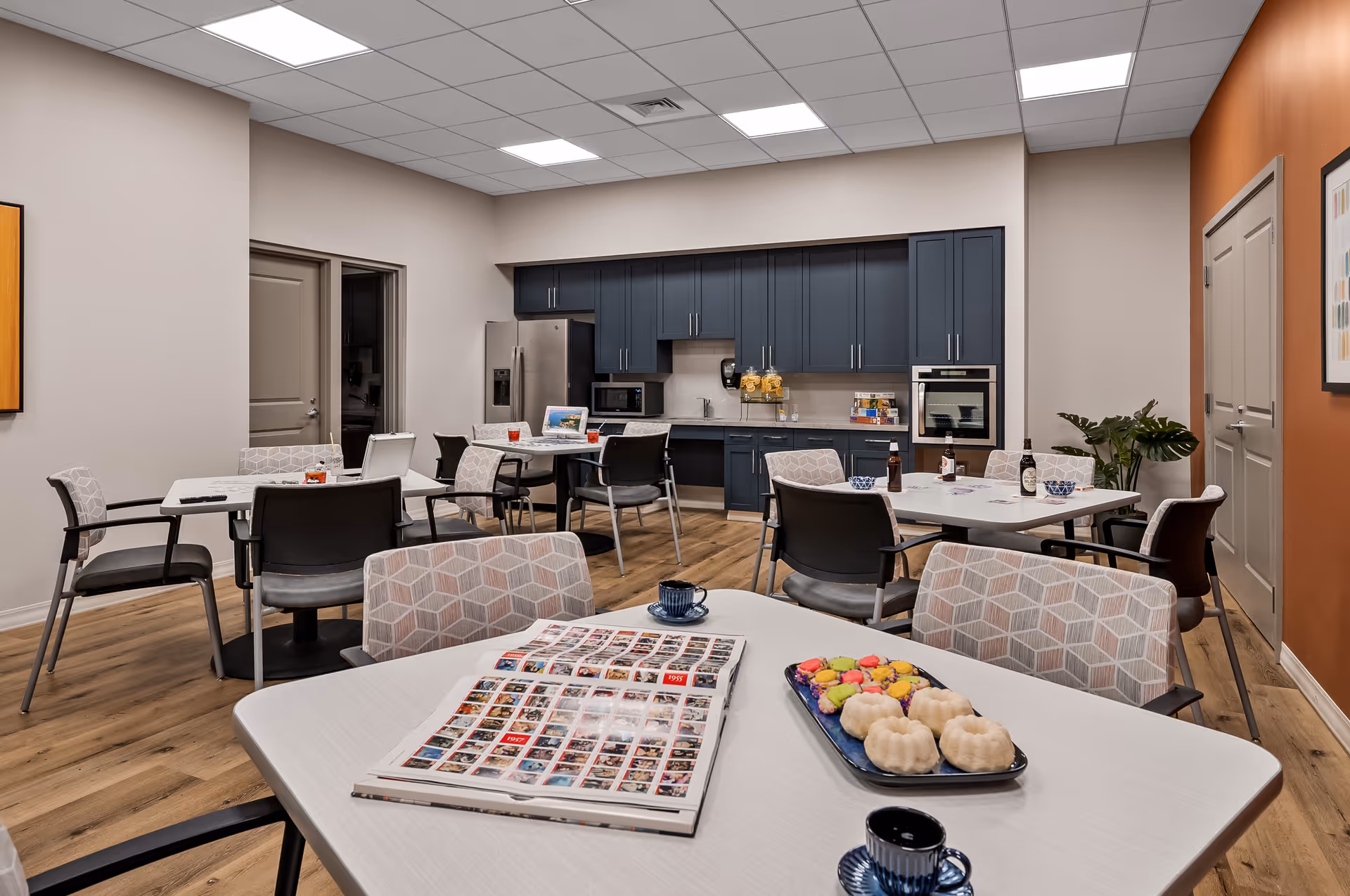 Communal dining area with multiple tables and chairs, a kitchenette with dark cabinets and stainless appliances, and snacks on the tables.