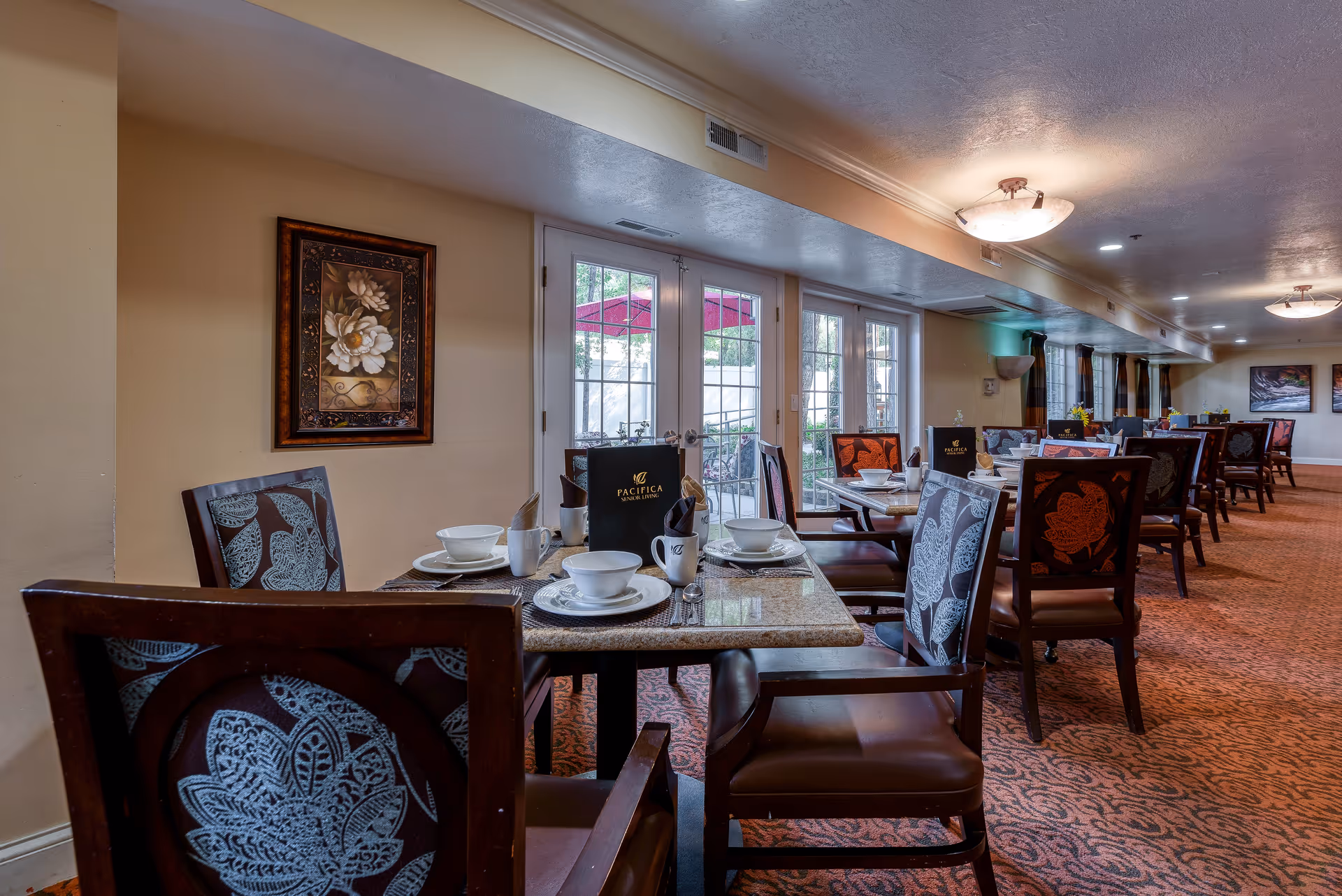 Interior view of a dining room with multiple tables set with white bowls, plates, cups, and silverware. The chairs have dark wooden frames with patterned upholstery. The room has beige walls, carpeted floor with a red and orange pattern, and large windows with glass doors leading outside. Wall art and ceiling lights are visible.