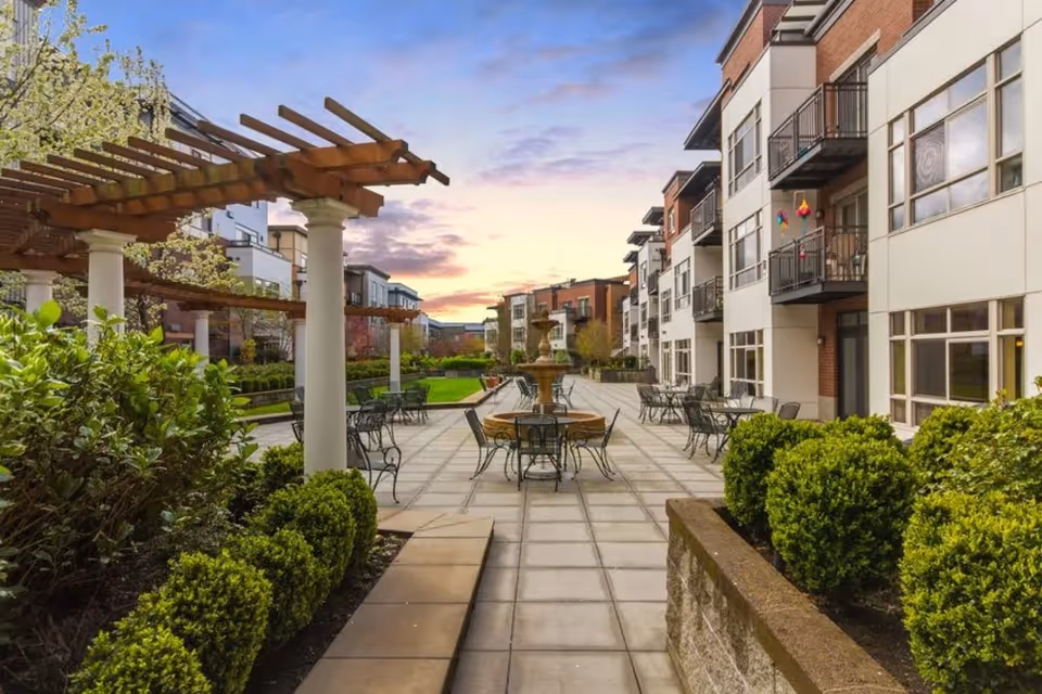 Outdoor patio area at Merrill Gardens at The University featuring a paved walkway with metal tables and chairs, a central water fountain, pergolas with wooden beams supported by white columns, and surrounding greenery and bushes. Apartment buildings with balconies line the right side under a partly cloudy sky at sunset.