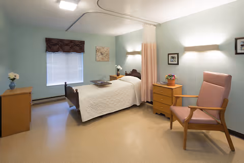 A clean and simple nursing facility room with a single bed covered in a white quilt, a wooden nightstand with a flower pot, a wooden dresser with a vase of flowers, a pink cushioned armchair, and a window with a valance. The walls are painted light green and there is a privacy curtain partially drawn around the bed.