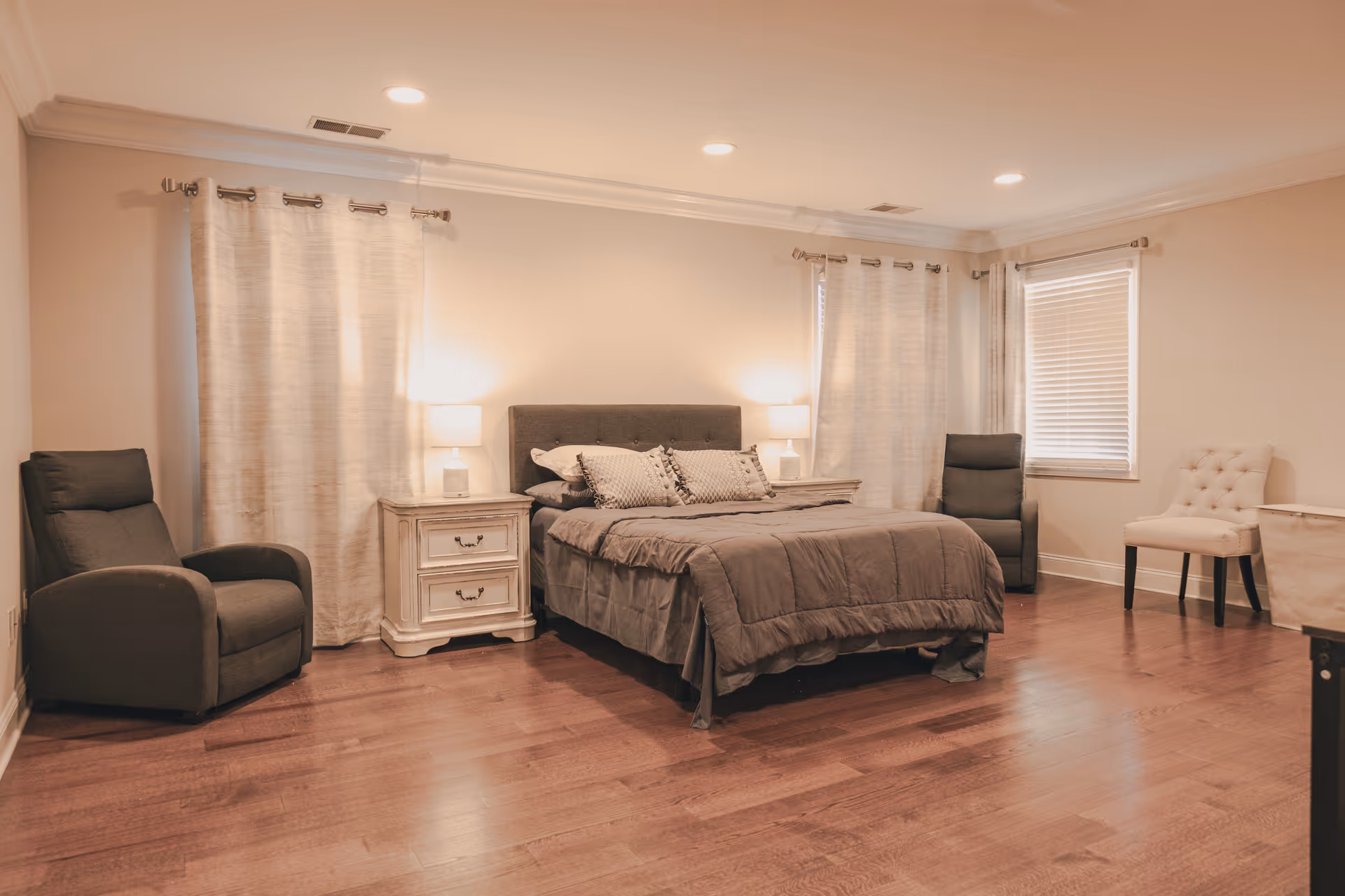 A spacious bedroom with a large bed in the center, flanked by two nightstands each with a lamp. There are two dark armchairs on either side of the bed, light-colored curtains on the windows, and a light-colored upholstered chair near the window. The room has wooden flooring and recessed ceiling lights.