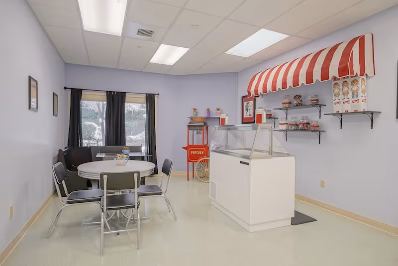 Small common area with a round table and chairs, a popcorn cart and a concession-style snack counter under a red-and-white striped awning.