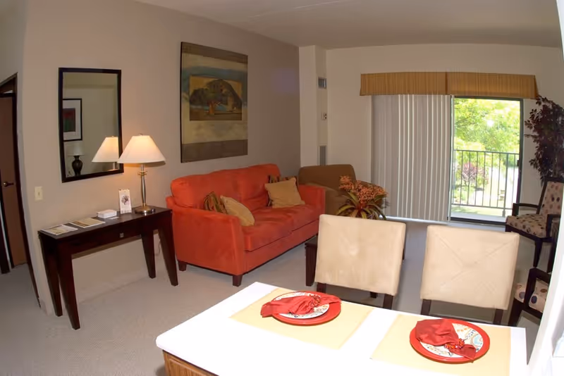 A cozy living room area with a red sofa and beige armchair, a wooden side table with a lamp and mirror above it, a painting on the wall, and a sliding glass door with vertical blinds leading to a balcony with greenery outside. In the foreground, there is a dining table set with two place settings, each with a red plate and folded red napkin on a beige placemat.