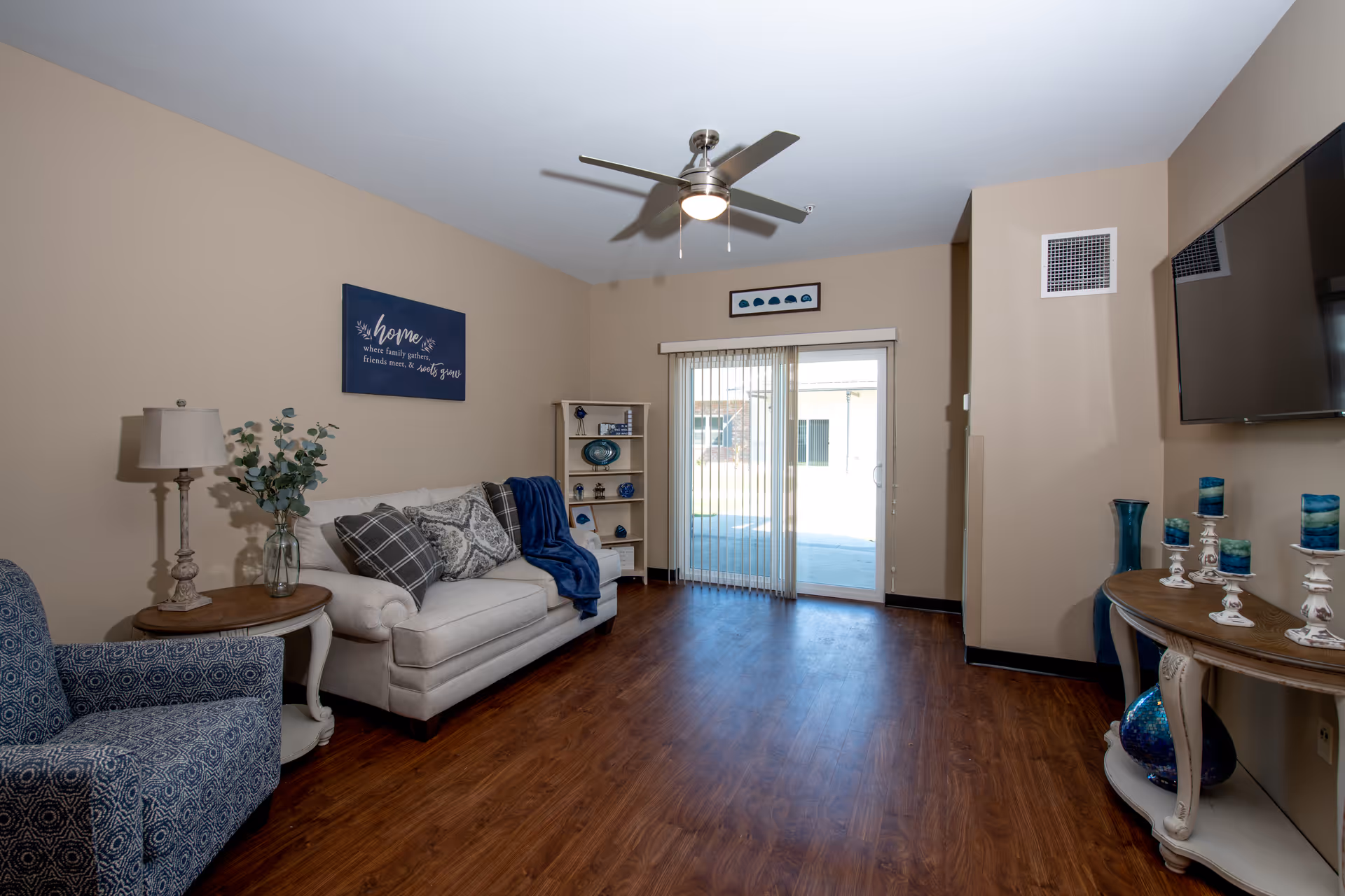 A cozy living room with beige walls and wooden flooring featuring a white sofa with patterned cushions and a blue throw, a blue patterned armchair, a small round wooden side table with a lamp and a vase with greenery, a white shelving unit with decorative items, a ceiling fan with light, a wall-mounted flat screen TV, and a console table with blue and white decorative candles and vases. Sliding glass doors with vertical blinds lead to an outdoor area.