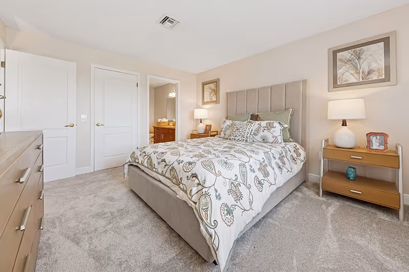 Well-lit bedroom with a upholstered bed dressed in patterned bedding, nightstands with lamps, a dresser, and a doorway to an en-suite bathroom.
