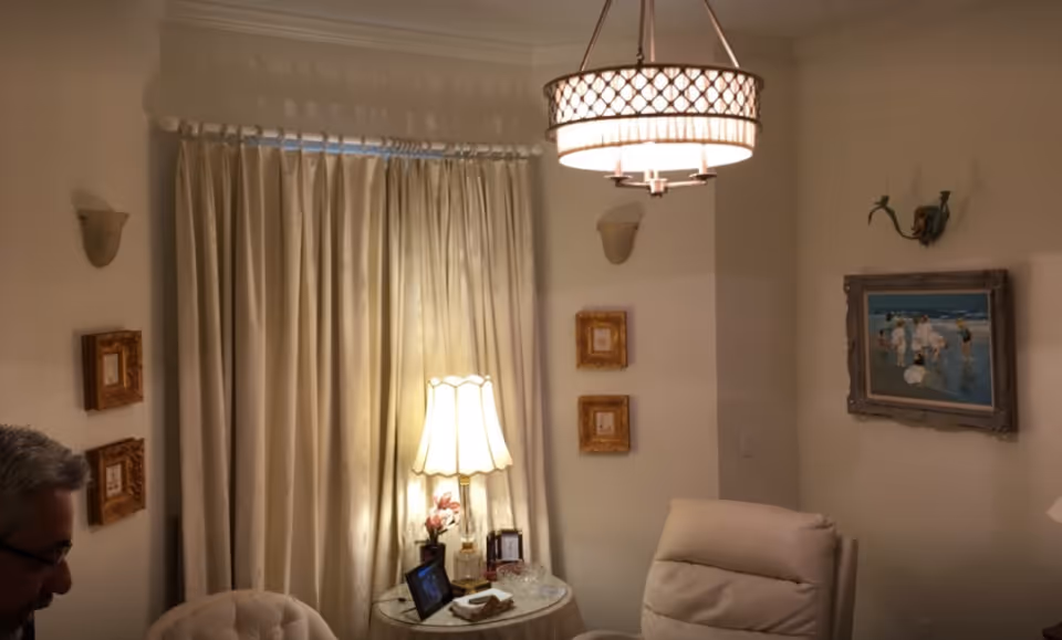 A cozy living room area with cream-colored walls and curtains. There is a round side table with a lamp, a small flower arrangement, and some framed photos. Two comfortable chairs are visible, one tufted and one recliner. The room is decorated with framed artwork on the walls and a ceiling light fixture with a decorative shade.