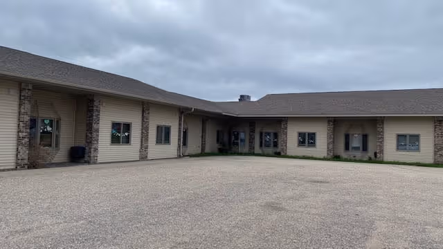 Exterior view of a single-story assisted living facility building with beige siding and brick accents under a cloudy sky. The building forms an L-shape around a large paved area in front.