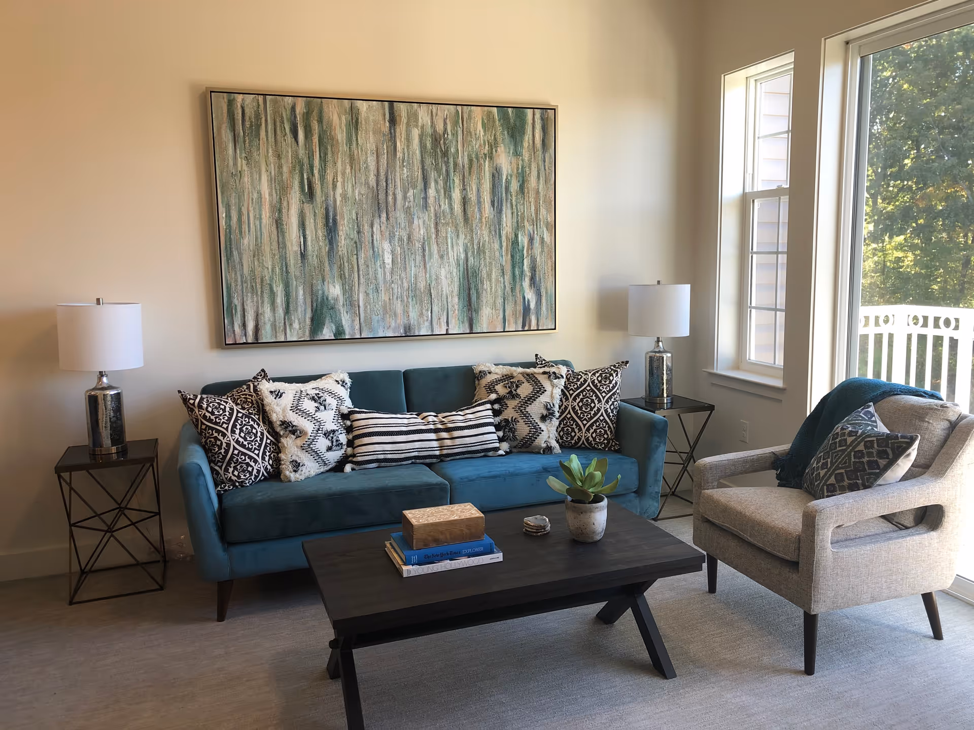A cozy living room with a teal sofa adorned with patterned throw pillows, a beige armchair with a dark blue throw and pillow, a dark wooden coffee table with books, a small plant, and decorative items. Two side tables with matching lamps flank the sofa. A large abstract painting hangs on the wall above the sofa, and a window on the right lets in natural light, showing greenery outside.