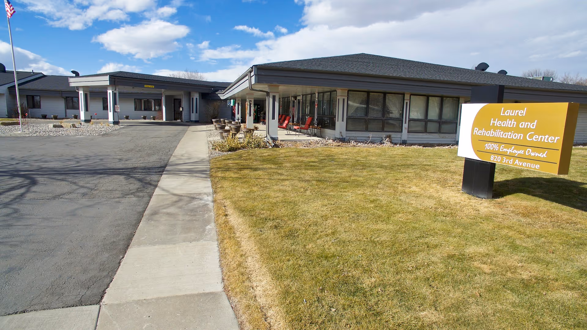 Exterior view of Laurel Health and Rehabilitation Center showing a single-story building with a paved driveway, a sidewalk, a grassy lawn, and a sign with the facility's name and address. The sky is partly cloudy.