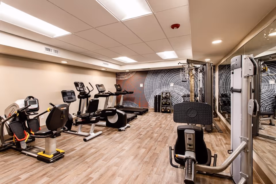 A fitness room with various exercise equipment including stationary bikes, treadmills, and weight machines. The room has wood-patterned flooring, a mirrored wall on the right, and a decorative wall with circular patterns at the far end.