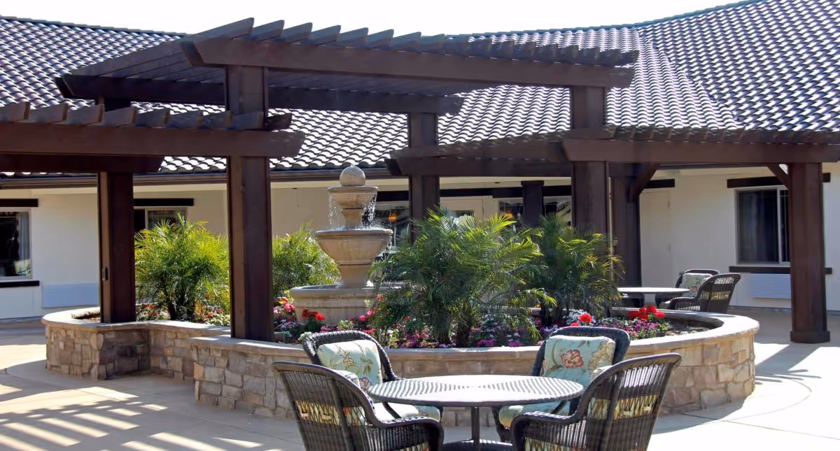 Outdoor courtyard area with a stone planter filled with green plants and colorful flowers surrounding a tiered water fountain. There are wooden pergolas providing partial shade and several wicker chairs with floral cushions around round tables on a paved surface. The building with a tiled roof and windows is visible in the background.