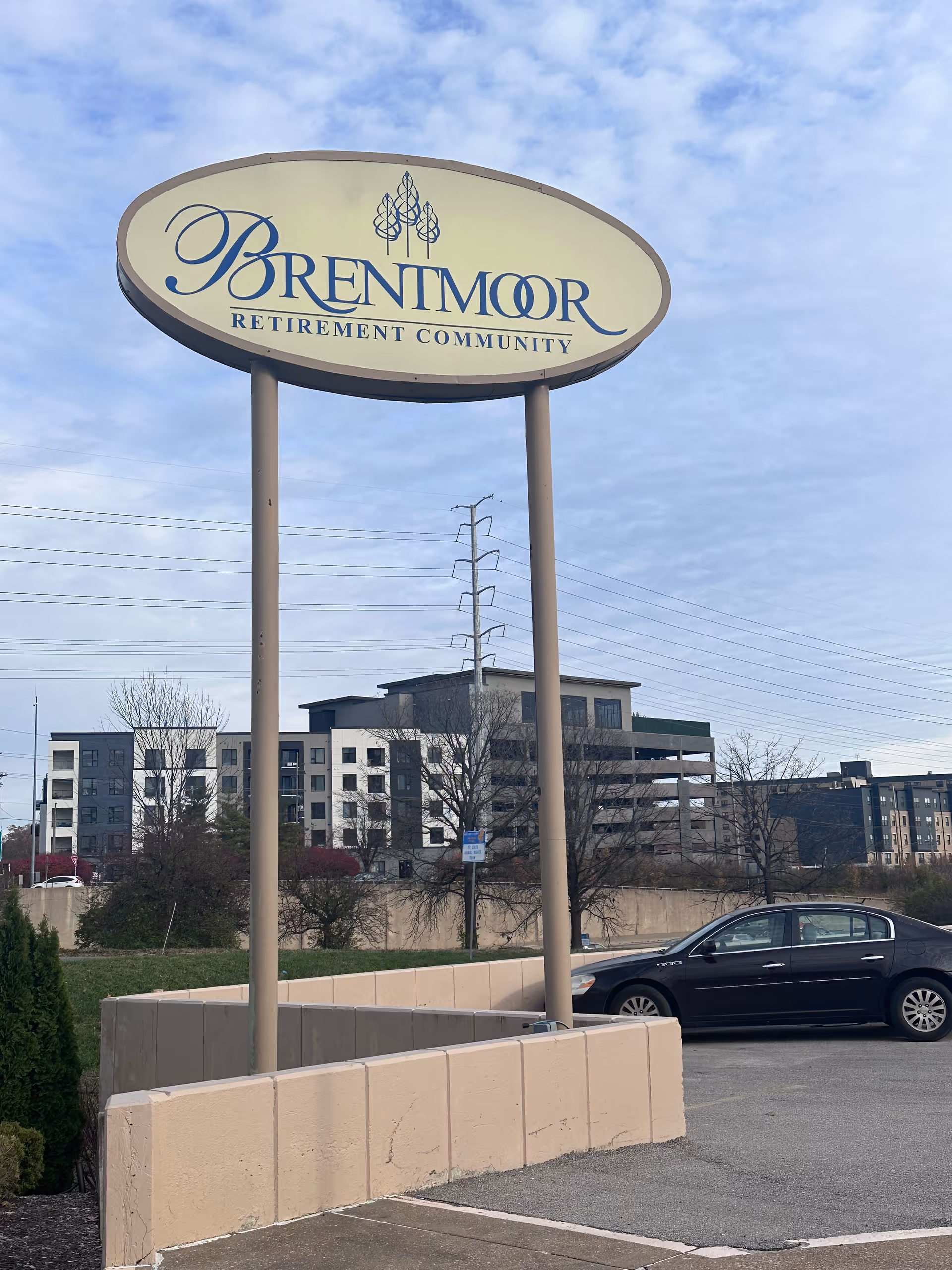 A large oval sign for Brentmoor Retirement Community mounted on two tall poles in a parking lot. Behind the sign, there are trees, a black car parked, and multi-story buildings under a partly cloudy sky.