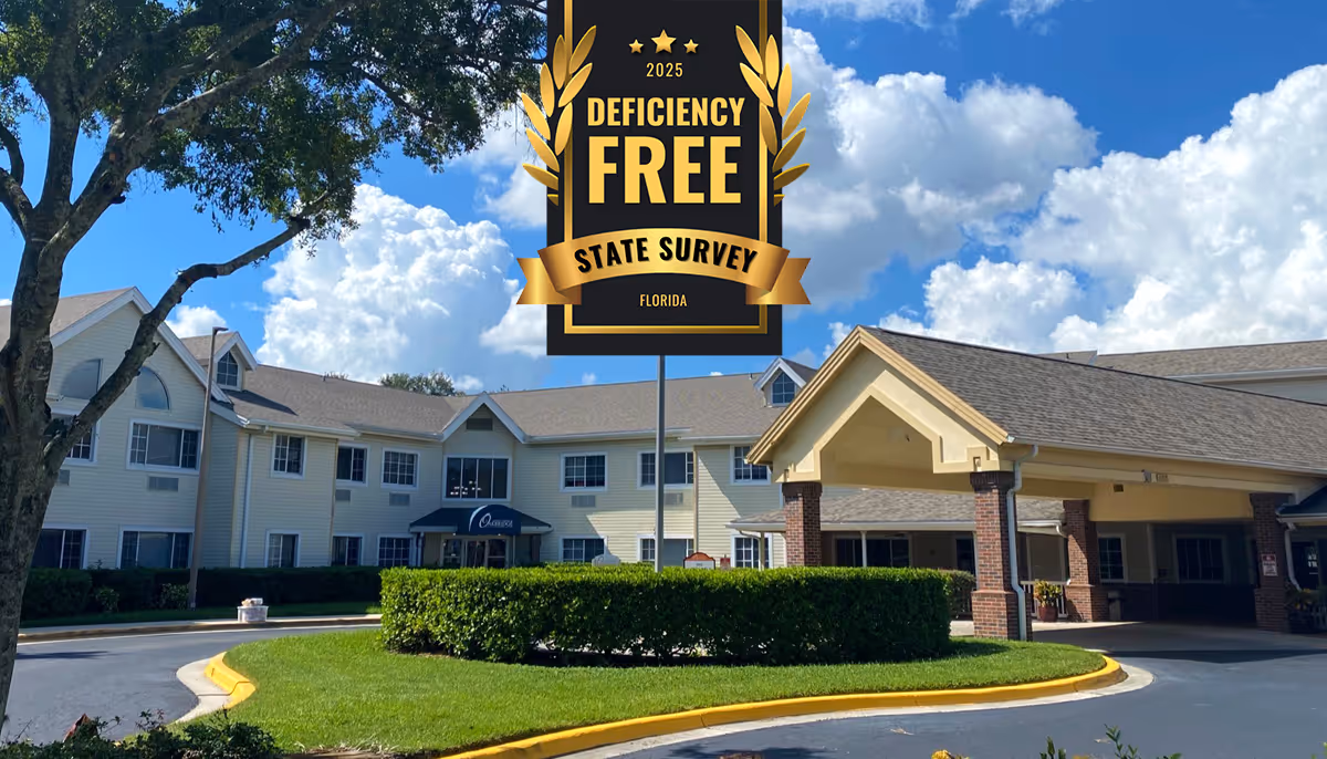 Exterior view of a senior living facility building with a covered entrance and well-maintained landscaping under a partly cloudy blue sky. A large tree is visible on the left side of the image.