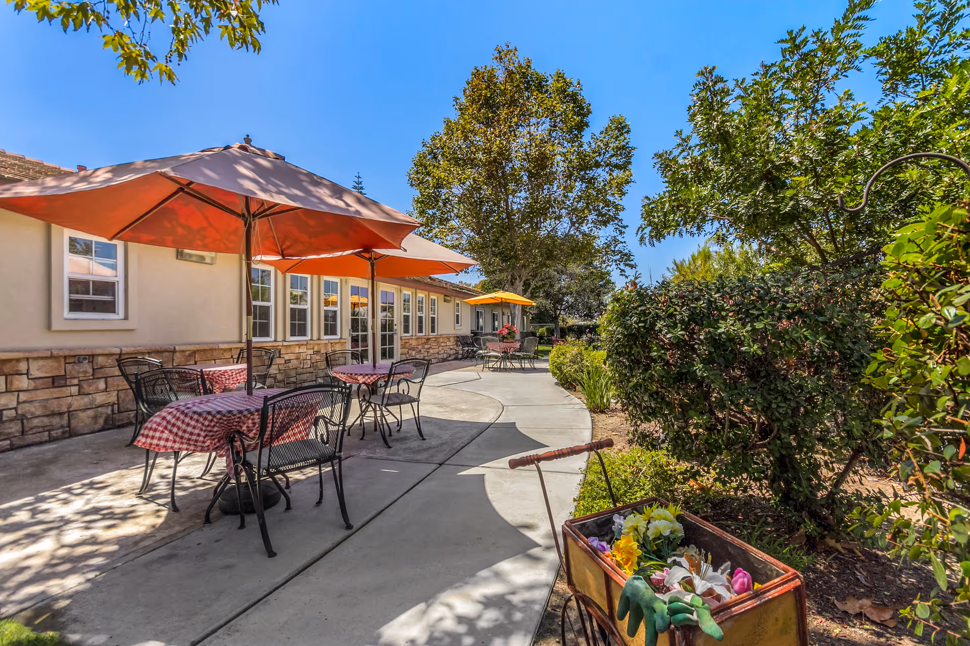 Outdoor patio area with several tables covered with red checkered tablecloths and large umbrellas providing shade. The patio is adjacent to a building with stone and stucco exterior walls and multiple windows. There are trees and bushes surrounding the patio, and a small cart filled with gardening gloves and flowers is visible in the foreground.