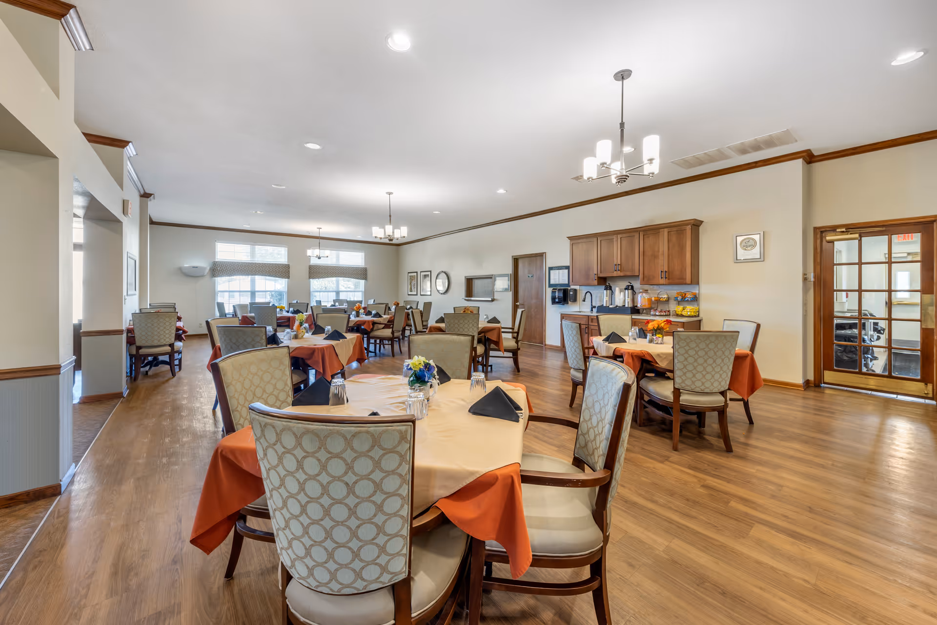 A spacious dining room with multiple round tables covered with beige and orange tablecloths, each set with black napkins and glassware. The room has wooden flooring, beige walls with wooden trim, and several chandeliers hanging from the ceiling. There is a counter with cabinets, coffee dispensers, and snacks along one wall, and large windows letting in natural light.