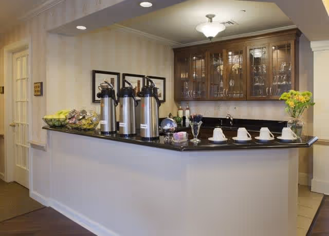 A coffee station area with a black countertop featuring three stainless steel coffee dispensers, a basket of bananas and other fruit, four white coffee cups on saucers, and a vase with yellow flowers. Behind the counter are wooden cabinets with glass doors displaying glassware, and a wall with framed pictures. The area is well-lit with a ceiling light fixture.