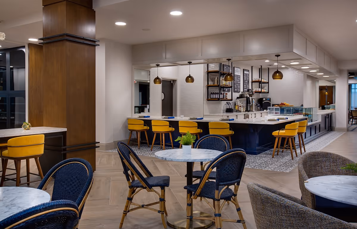 Interior view of a modern dining and lounge area with marble-top tables, blue woven chairs, and yellow bar stools at a counter with pendant lighting. The space features wood flooring, a wooden column, and a coffee or snack bar area in the background.