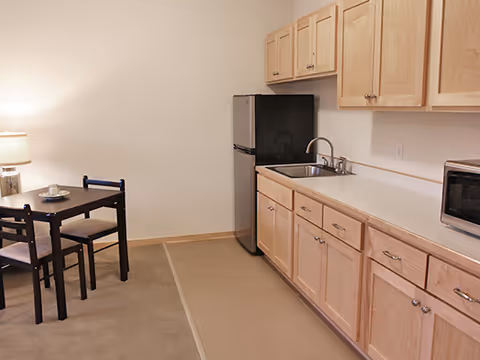 Small kitchenette with light wood cabinets, a sink, mini-fridge and microwave, and a small dining table with two chairs.