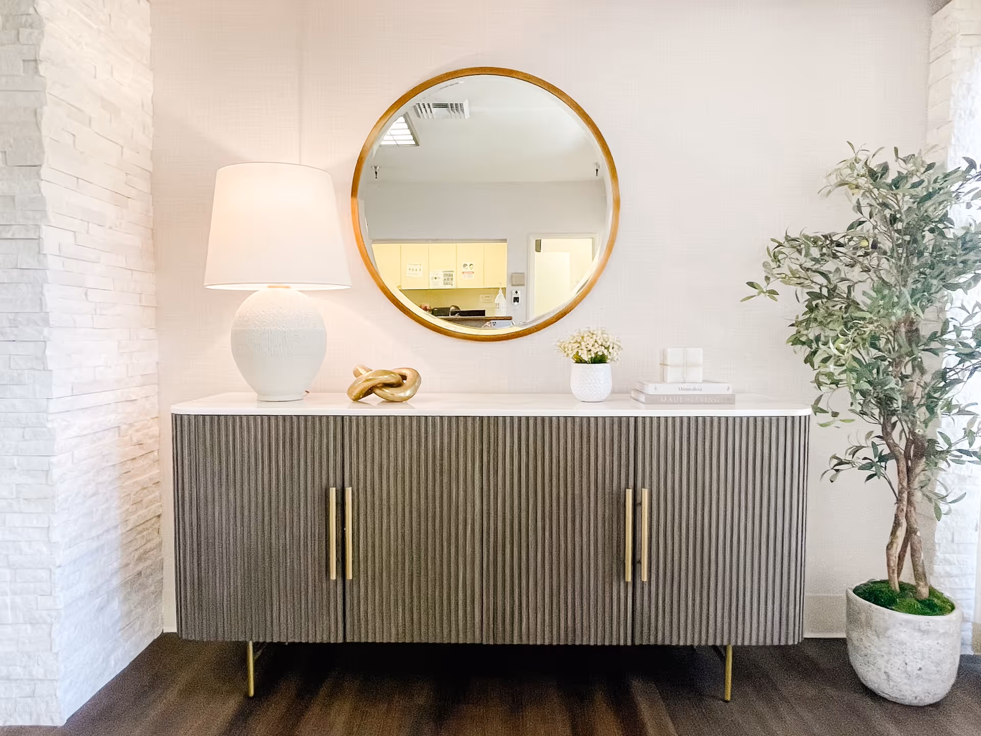 A modern interior corner featuring a textured gray cabinet with gold handles, topped with a white lamp, a gold decorative knot, a small white vase with flowers, and stacked books. Above the cabinet is a round mirror with a wooden frame reflecting part of the room. To the right of the cabinet is a tall potted plant with green leaves. The walls are light-colored with a textured white brick accent on the left side and dark wood flooring.