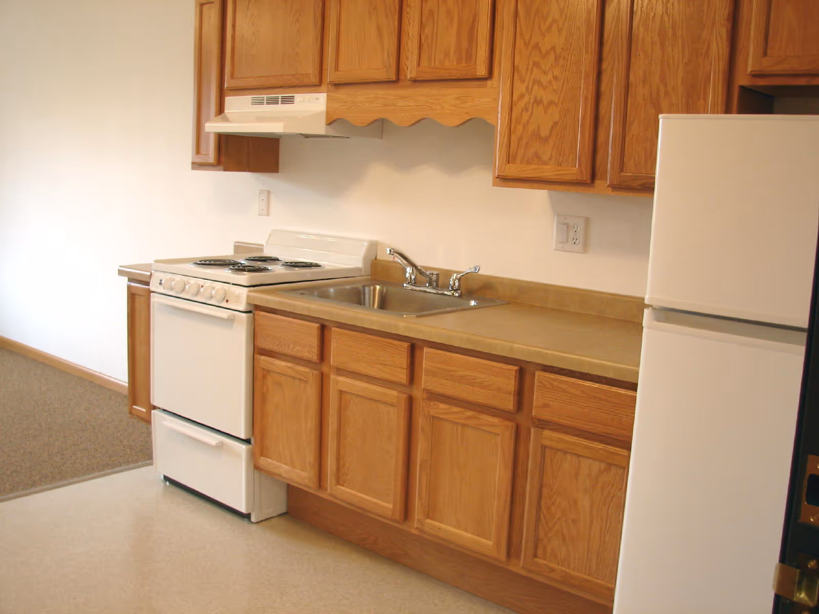 A small kitchen area with wooden cabinets, a white electric stove with four burners, a stainless steel sink with a faucet, and a white refrigerator. The kitchen has a beige countertop and light-colored flooring, with a carpeted area visible in the background.