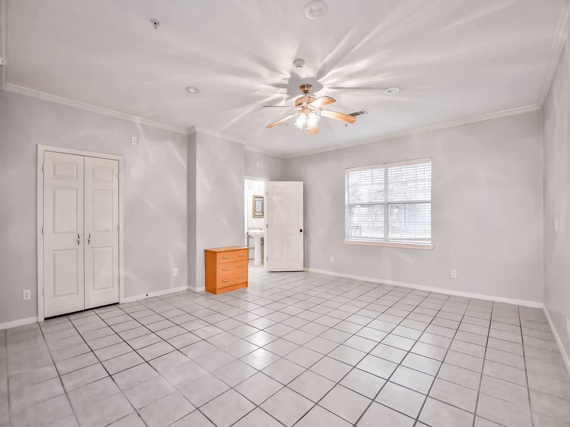 An empty room with light gray walls and white tiled floor. There is a ceiling fan with lights in the center of the ceiling. A window with white blinds is on the right wall, and a small orange dresser is against the far wall near an open door leading to a bathroom. A closed double-door closet is on the left side of the room.
