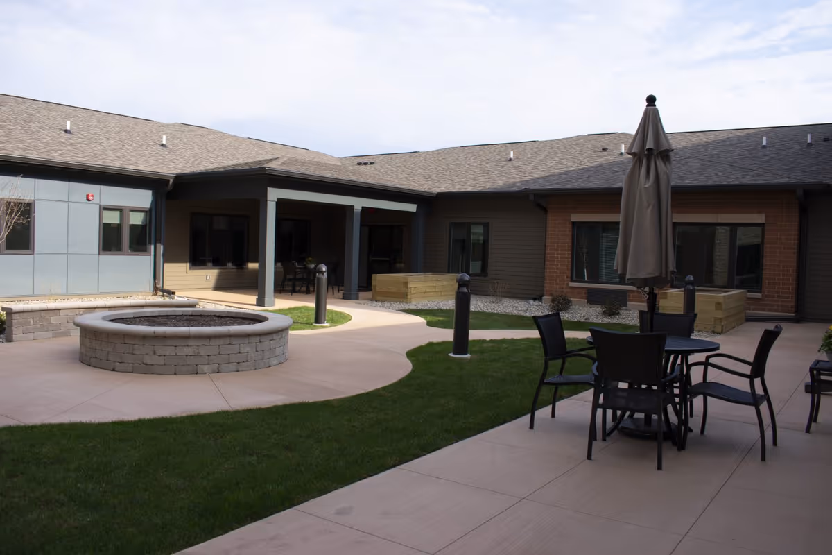 Courtyard with patio seating and umbrella, a circular stone firepit, and surrounding single-story facility building.