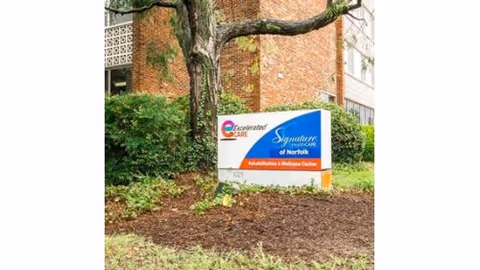 Outdoor view of a sign for Signature HealthCARE of Norfolk, a Rehabilitation & Wellness Center, placed in a landscaped area with mulch, grass, bushes, and a tree. A brick building is partially visible in the background.