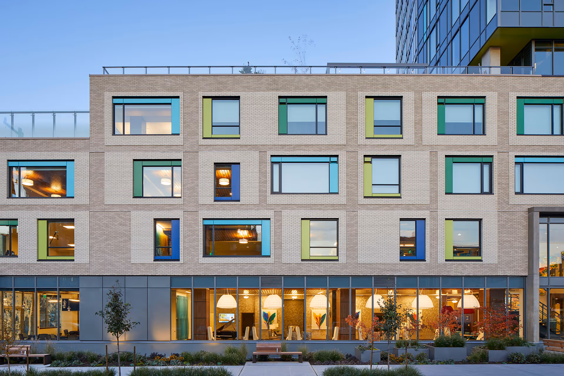 Exterior view of a modern multi-story building with large windows, some with colorful frames in blue, green, and yellow. The ground floor features large glass windows revealing a warmly lit interior with hanging pendant lights and seating areas. There are small trees and landscaped greenery in front of the building under a clear sky.