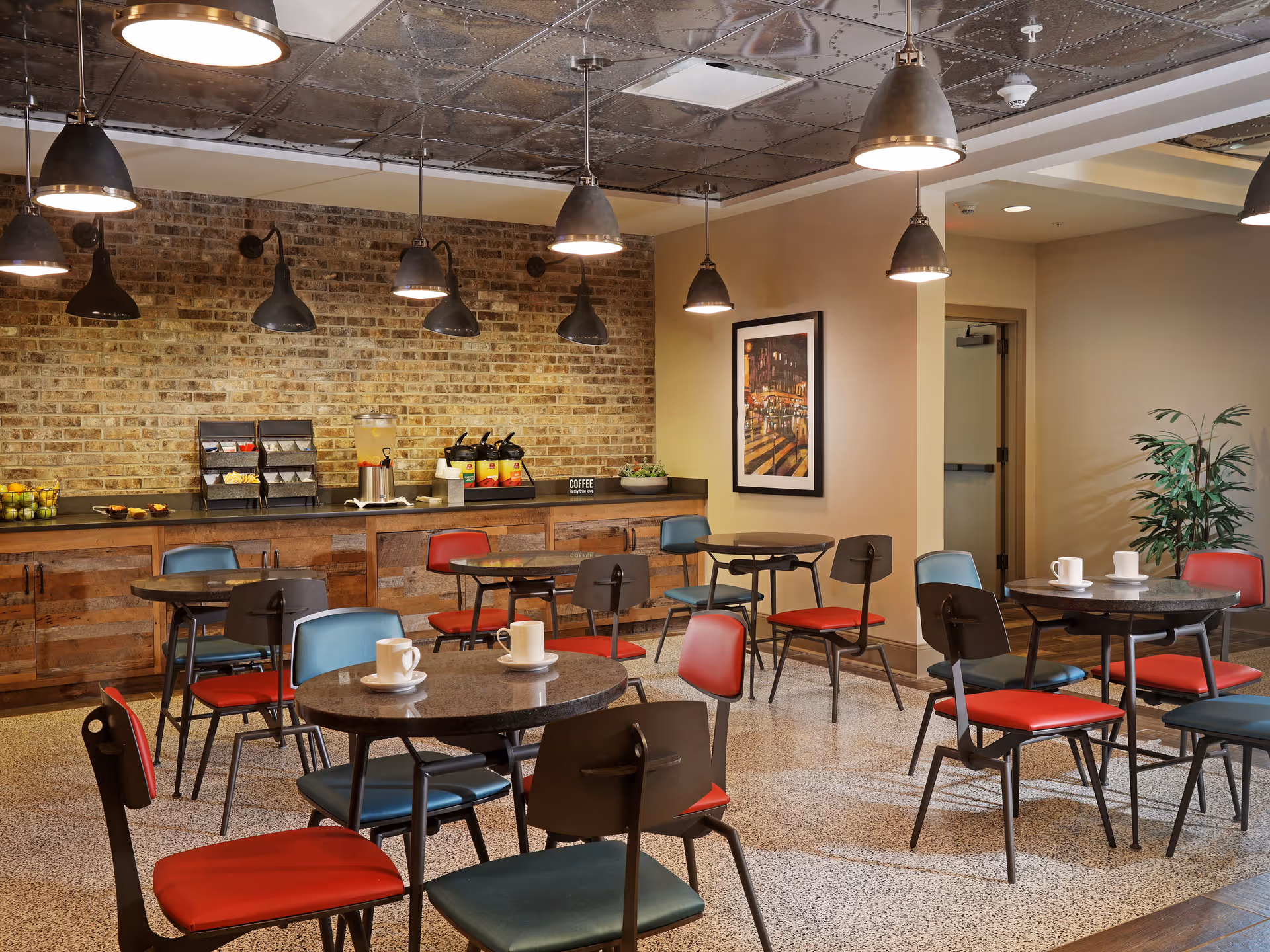 A brightly lit dining room with round tables and red and blue chairs, pendant lights, and a brick back wall with a beverage station.