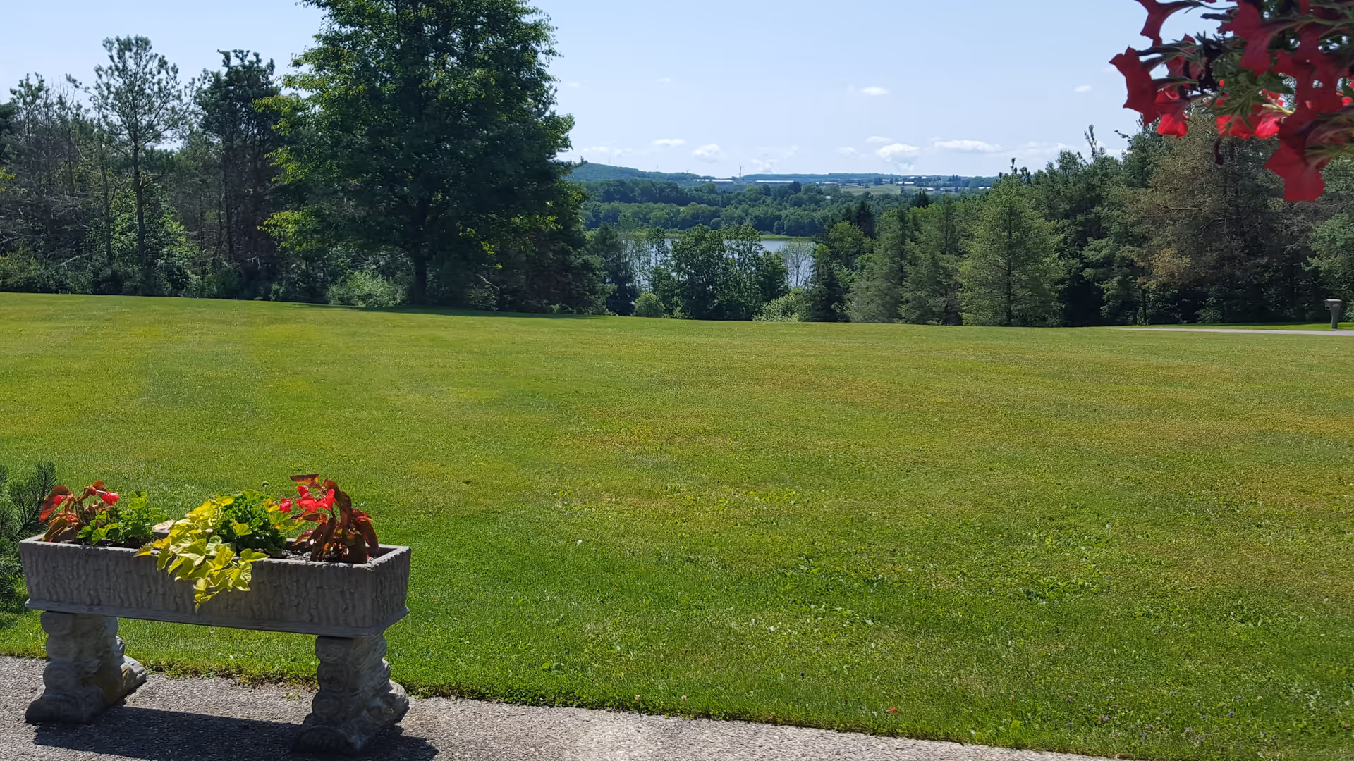 A wide green lawn with a stone planter filled with colorful flowers in the foreground. Trees surround the lawn, and a body of water is visible in the distance under a clear blue sky with a few clouds.