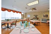 A dining room with several tables covered in light pink tablecloths, set with plates, glasses, and silverware. The room has large windows with sheer white curtains and orange valances, wooden flooring, and floral wallpaper. There are chairs around the tables and decorative items on the walls and a sideboard.