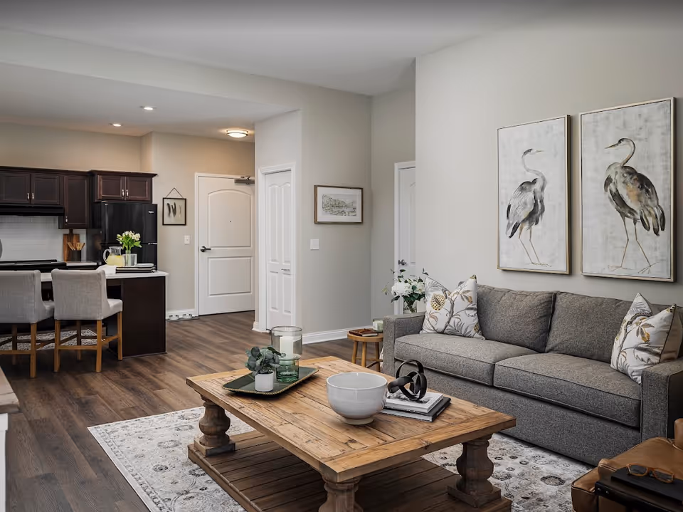 A modern living room and kitchen area in an active adult community. The living room features a gray sofa with patterned pillows, two framed bird artworks on the wall, a wooden coffee table with decorative items, and a patterned rug. The kitchen has dark wood cabinets, a black refrigerator, a white countertop with two gray upholstered bar stools, and a vase with flowers. The flooring is wood, and there are two white doors visible in the background.