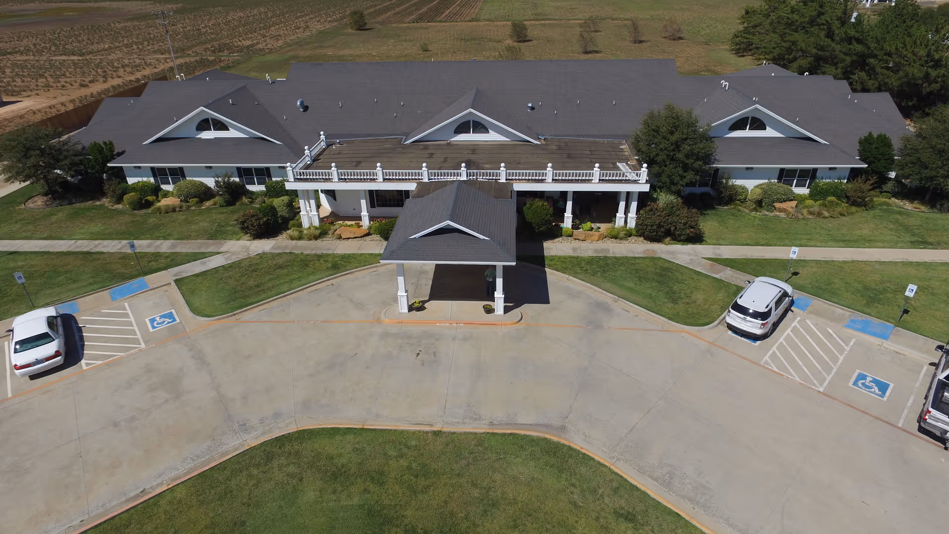 Aerial view of the building front showing a single-story senior living facility with a covered entrance drive, parked cars, handicap spaces, and lawns.