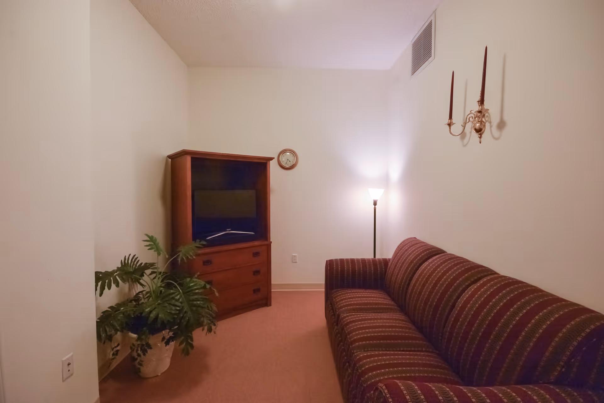 A small living room with a striped maroon sofa on the right, a wooden TV cabinet with a television in the corner, a potted plant on the floor to the left, a floor lamp in the back corner, a small round wall clock above the TV cabinet, and a decorative wall sconce with two candles on the right wall.