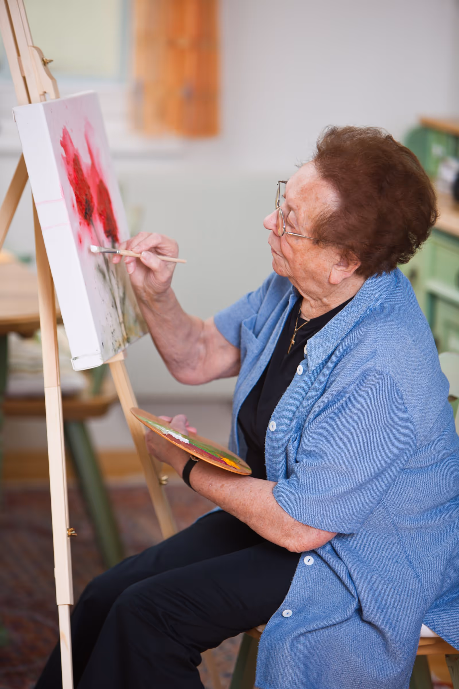 An elderly woman wearing glasses and a blue shirt is sitting and painting on a canvas placed on an easel. She holds a paint palette in one hand and a paintbrush in the other, applying red and black paint to the canvas. The setting appears to be indoors with furniture and a window in the background.
