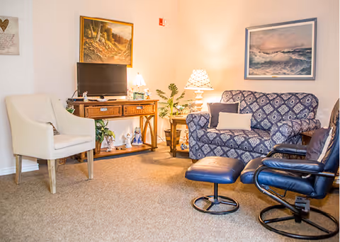 A cozy living room area with a patterned blue loveseat, a black leather swivel chair with matching ottoman, a white armchair, and a wooden TV stand holding a flat screen TV and decorative items. Two table lamps provide warm lighting, and framed artwork hangs on the walls.