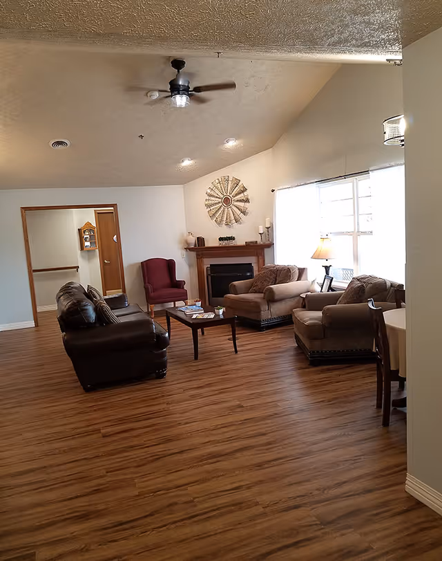 Spacious senior living common room with sofas and armchairs arranged around a coffee table and fireplace beneath a large window.