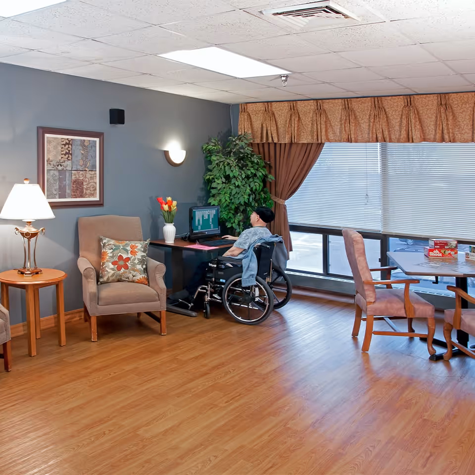 A bright senior living lounge with chairs and tables where a person in a wheelchair uses a computer by a window.
