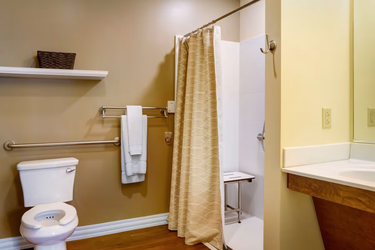 Bathroom with a white toilet, a towel rack holding two white towels, a shower area with a beige patterned curtain and a white shower chair inside, a wall-mounted grab bar, a shelf with a basket, and a sink with a large mirror.