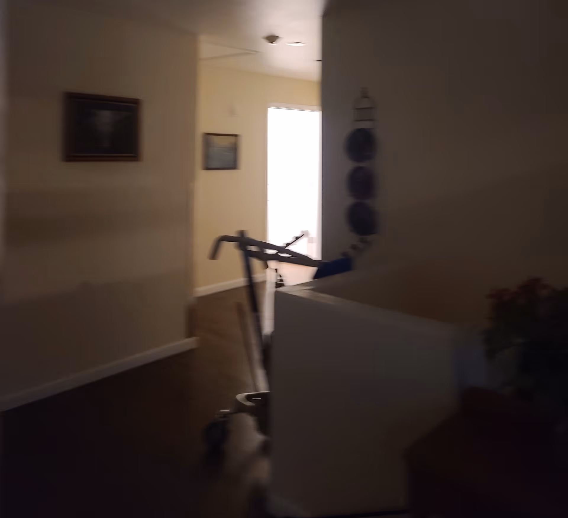 Dimly lit interior hallway of a facility with a walker positioned near a wall. The hallway leads to a brighter room with framed pictures on the walls and wooden flooring.