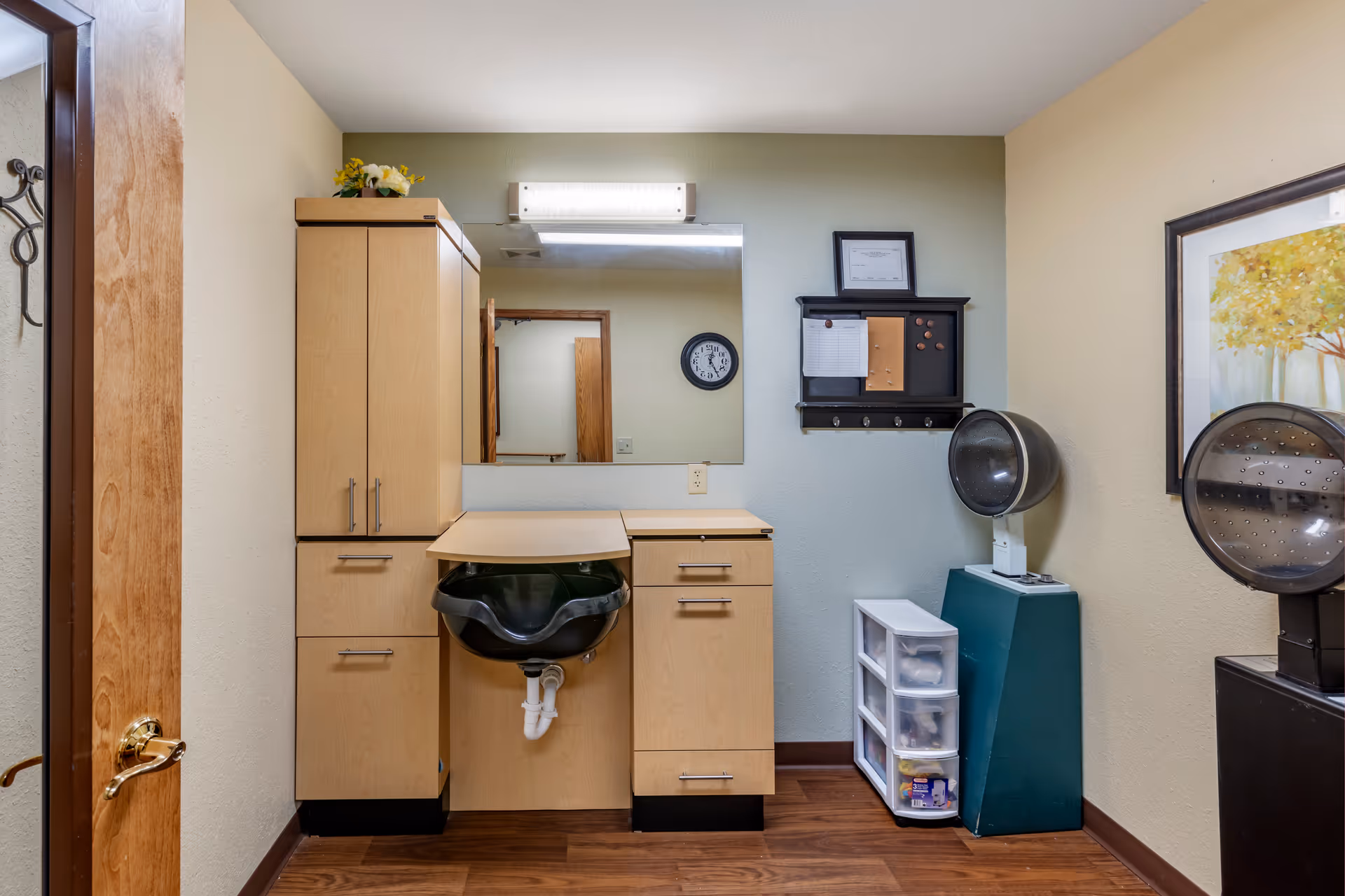 Interior room with a wooden floor and beige walls, featuring a light wood cabinet with drawers and a black sink. Above the sink is a large mirror with a light fixture. To the right, there is a black hair dryer chair and a small plastic drawer unit. A framed picture of trees hangs on the wall.