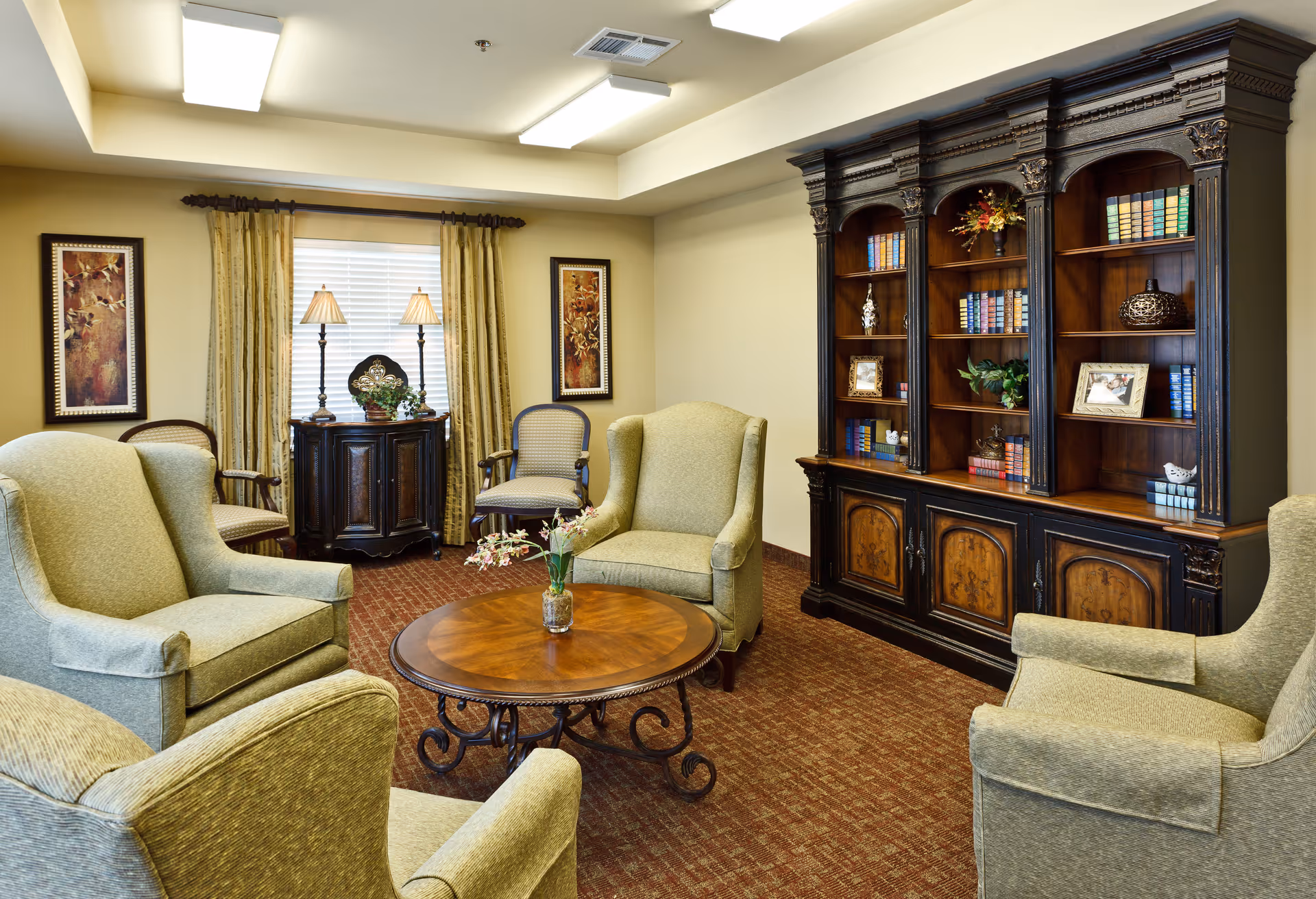 A cozy living room with four green upholstered armchairs arranged around a round wooden coffee table with a small flower vase. Behind the chairs is a large dark wooden bookshelf filled with books, framed photos, and decorative items. The room has beige walls, carpeted floor, two framed paintings, a window with curtains, and a small cabinet with two lamps on top.
