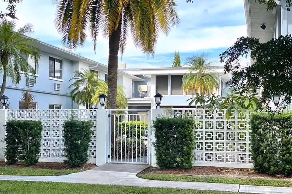Exterior view of a senior living facility named Elite at Cape Coral featuring a white decorative concrete fence with a metal gate, trimmed bushes, palm trees, and a two-story building with balconies and windows in the background under a partly cloudy sky.
