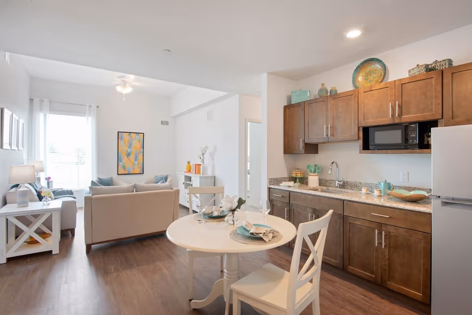 Bright open-concept living area and kitchenette with a round dining table, beige sofa, and wood cabinets.